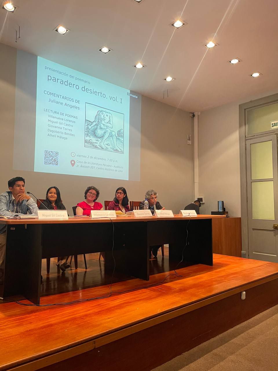 Foto grupal: Villanueva Lorenzo, Giovanna Torres, Alhelí Málaga, Julianne Ángeles y Dagoberto Benites sentados en la mesa de presentación, detrás de ellos, en la pared, se proyecta el afiche que convoca a la presentación del libro